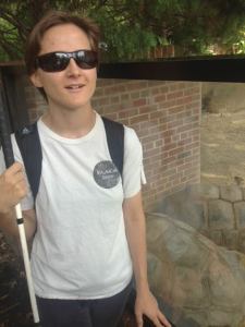 Young white person with a white cane smiling outdoors in front of brick walls and a glass partition with large tortoises at the zoo.